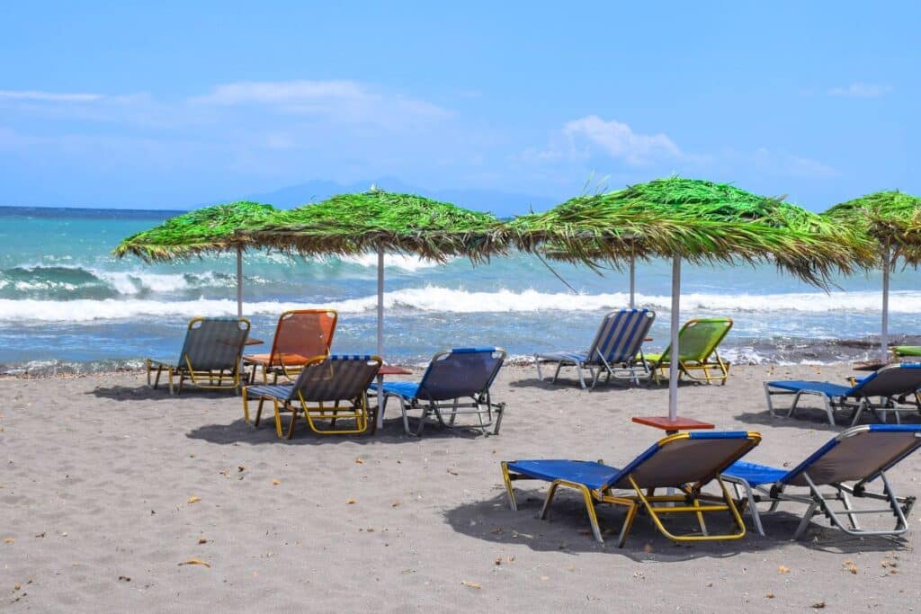 A black sand beach in Santorini dotted with colorful lounge chairs and green thatched umbrellas